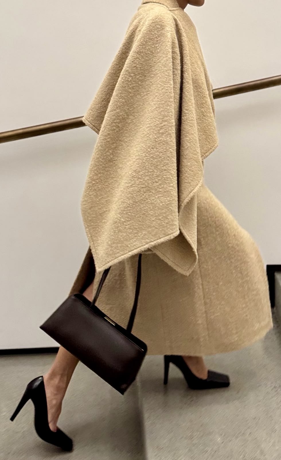 Person i en beige kappa med struktur, mörkbrun handväska och svarta skor på väg uppför en trappa.
