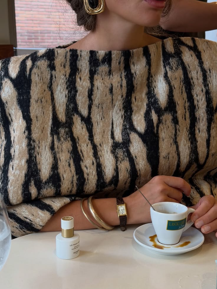 Närbild av en person i en beige och svart mönstrad topp med smycken och en espressokopp vid ett bord.