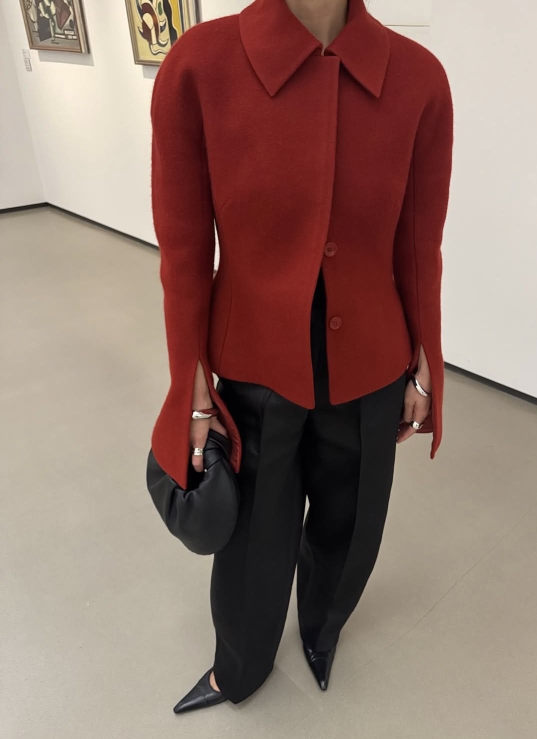 Person i röd figursydd jacka, svarta vida byxor och spetsiga skor i en gallerimiljö.