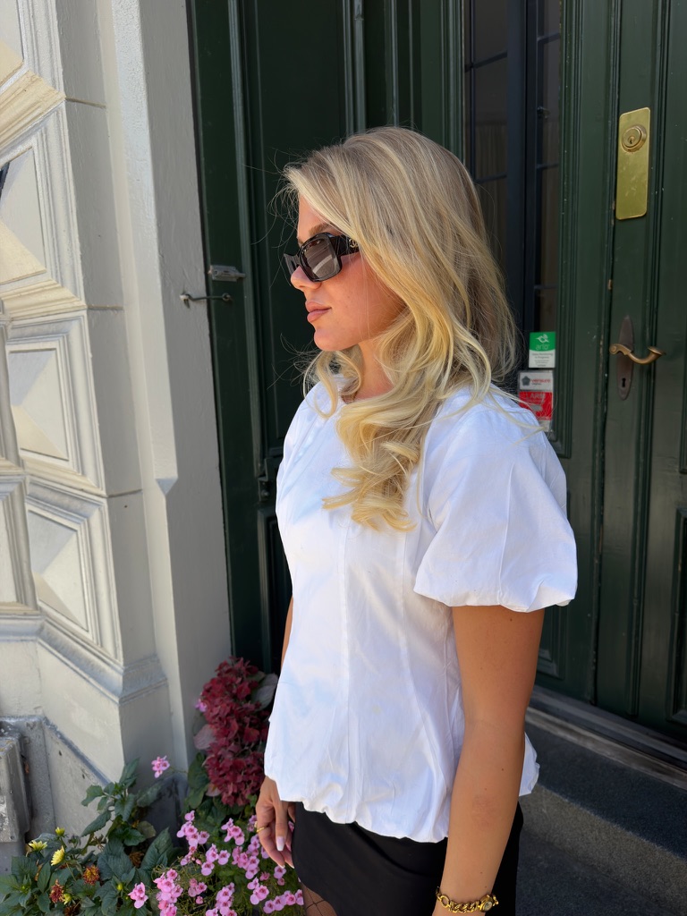 Person i vit puffärmad topp och solglasögon framför en mörkgrön dörr.
